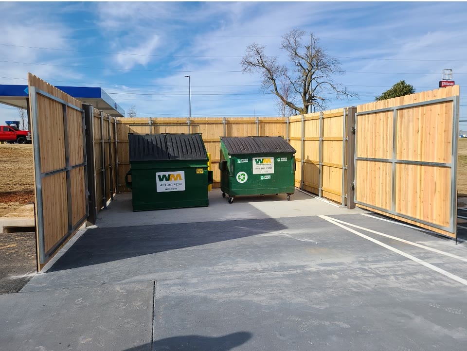 Cedar-and-steel dumpster enclosure with two swing gates open at a commercial lot in Northwest Arkansas