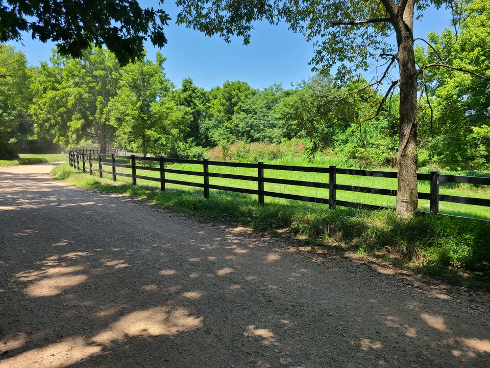 Black vinyl ranch-rail fence running along a tree-lined gravel drive in Northwest Arkansas